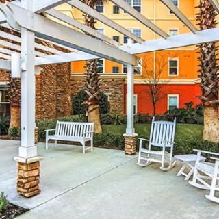 A cozy outdoor seating area with benches and rocking chairs under a pergola, surrounded by lush landscaping and palm trees.