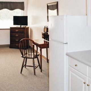 A cozy apartment interior with a wooden desk and chair, a dresser with a television, and a refrigerator.