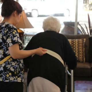 A caregiver assists an elderly person using a walker.