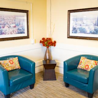 A cozy seating area featuring two teal armchairs with decorative pillows, a small table, and floral arrangements, complemented by framed artwork on the walls.