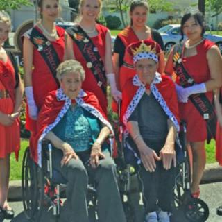A group of people posing outdoors, with two elderly individuals in wheelchairs wearing crowns and red capes, surrounded by younger people wearing red dresses and sashes.