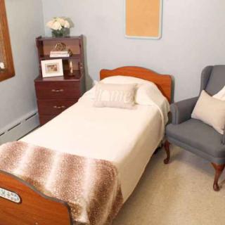 A cozy bedroom featuring a quaint single bed, an upholstered chair, and a wooden nightstand with decorative elements.