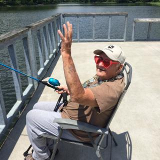 A man enjoying a sunny day fishing on a pier by the lake, wearing red sunglasses and a hat.