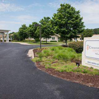 A paved road leading to a building, surrounded by trees and landscaped greenery, with a sign on the right.