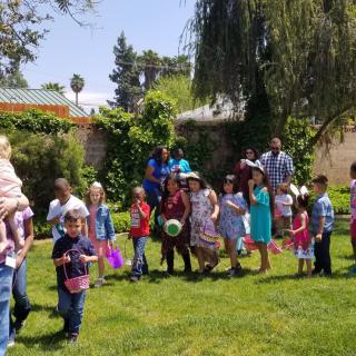A group of children and adults are gathered outdoors in a garden setting, enjoying a sunny day.