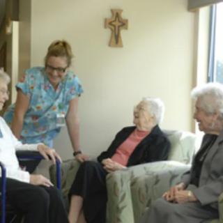 A group of elderly women in conversation with a caregiver in a nursing home setting.