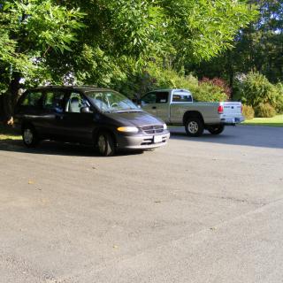 A minivan and a truck parked in a sunny parking lot surrounded by trees.