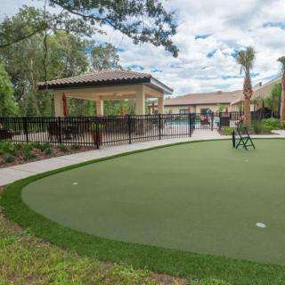 An outdoor putting green surrounded by trees and fencing, with a pavilion in the background.