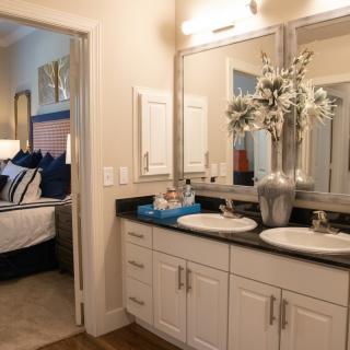 A modern bathroom with double sinks and mirrors, featuring a decorative vase with flowers and a view into the adjoining bedroom.