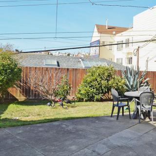 A sunny backyard with a patio area featuring a table and chairs, surrounded by greenery and enclosed by a wooden fence.