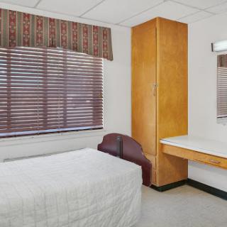 A simple and minimalistic bedroom featuring a bed, wooden furniture, and natural light from the window.