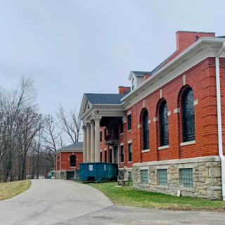 A historic red brick building set against a backdrop of nature, featuring large windows and a winding road.