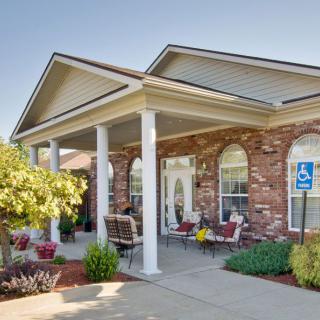 A welcoming building entrance with brick walls, white columns, and outdoor seating surrounded by landscaping.