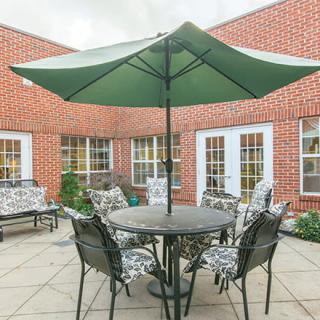 A cozy outdoor patio area featuring a table with chairs and a large green umbrella, surrounded by brick walls and windows.