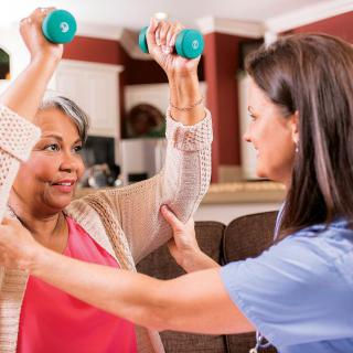 A therapist assists an elderly woman in exercising with dumbbells at home.