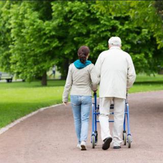 An elderly person using a walker, accompanied by a caregiver, walking in the park.