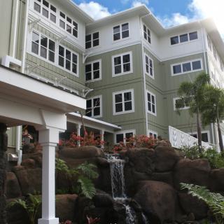 A modern green apartment building with multiple windows, surrounded by tropical landscaping and a cascading waterfall.
