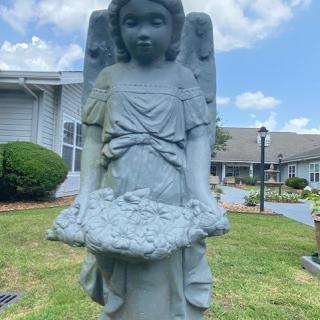 A stone angel statue in a garden setting with buildings in the background under a blue sky.