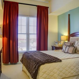 A cozy bedroom with a neatly made bed, adorned with pillows and a blanket, featuring red curtains and soft lighting.