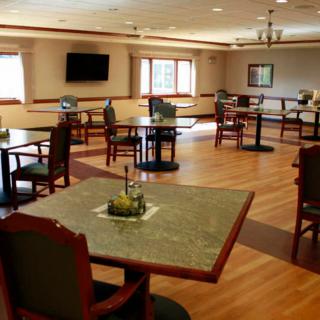 An empty dining room with tables and chairs arranged on a hardwood floor, featuring a television and large windows.
