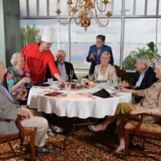 A group of seniors dining at a restaurant, being served by a chef in a formal setting.