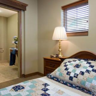 A cozy bedroom with a quilt-covered bed, a lamp on the nightstand, and a window with blinds.
