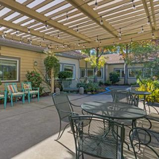 A cozy patio with metal furniture under a wooden pergola, surrounded by lush greenery.