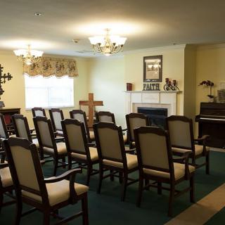 A small chapel interior with rows of chairs facing a cross and a piano.