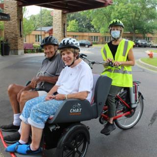 A volunteer riding a tricycle with two seniors seated in the front.