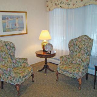 A cozy living room corner with two patterned armchairs, a small wooden table, and a lamp, set against light curtains.