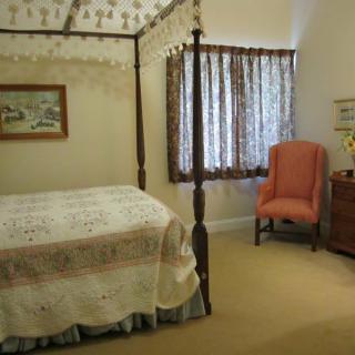 A cozy bedroom featuring a four-poster bed with a canopy and quilt, alongside an armchair, dresser, and window with curtains.