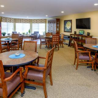 A spacious dining area with multiple tables and chairs, featuring large windows and a wall-mounted TV.