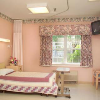 A hospital room featuring a bed, window with floral curtains, and a wall-mounted television.
