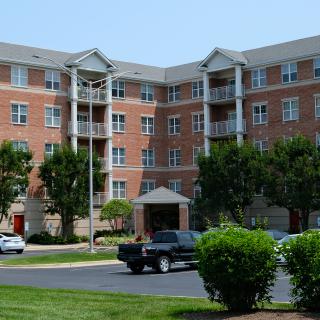 A multi-story brick apartment building with a parking lot and surrounding greenery.