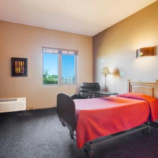 A cozy bedroom featuring a single bed with a red cover, a window allowing natural light, and comfortable furnishings including a chair and lamp.