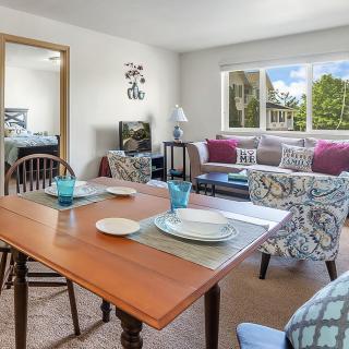 A cozy living and dining area with a wooden table set for two, comfortable seating, and a large window offering an outdoor view.