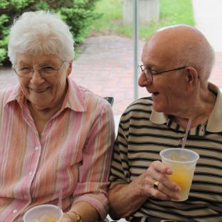 An elderly couple sitting together outdoors, enjoying drinks and smiling.