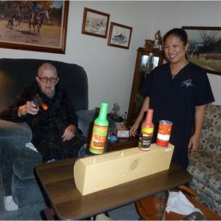 An elderly person sitting on a sofa next to a younger person standing, with various bottles placed on the table in front of them.