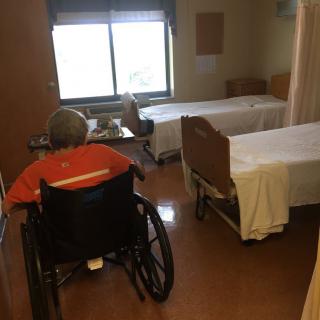 An elderly person in a wheelchair is sitting in a hospital room with two beds, near a window letting in daylight.
