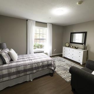 A modern, minimalist bedroom with a gray color scheme featuring a neatly made bed, a dresser with a mirror above it, and a window bringing in natural light.