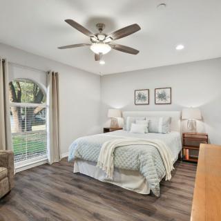 A cozy and modern bedroom with wood flooring, featuring a comfortable bed, nightstands with lamps, a window allowing natural light, a plush chair, and a ceiling fan for ventilation.