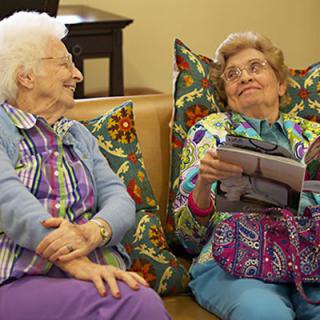 Two elderly women smiling and conversing while seated on a couch.