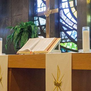 A church altar with an open Bible, candles, and a cross against stained glass windows.