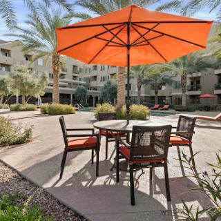 A cozy outdoor seating area features a bright orange umbrella providing shade over a round table and chairs, surrounded by landscaped greenery.