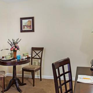 A cozy dining area with a small round table, adorned with flowers and surrounded by chairs, under a framed painting on the wall.