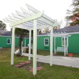 A white pergola stands in a yard with green buildings in the background, featuring a bench nearby.