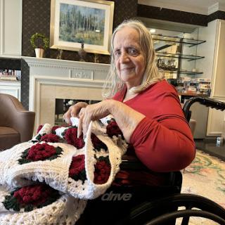An elderly woman in a wheelchair enjoys crocheting a beautiful blanket adorned with red roses.