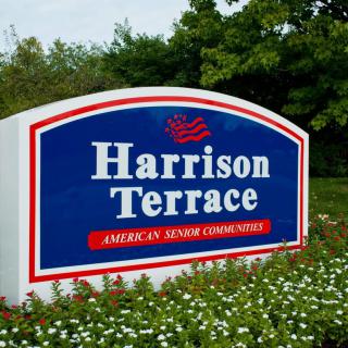 A sign for Harrison Terrace, part of American Senior Communities, surrounded by greenery and flowers.