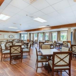 A spacious dining area with multiple tables and chairs, featuring wooden flooring and large windows with curtains.