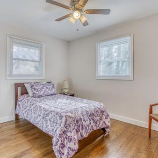 A cozy and well-lit bedroom featuring a single bed with a patterned cover, complemented by a ceiling fan and natural light from two windows.